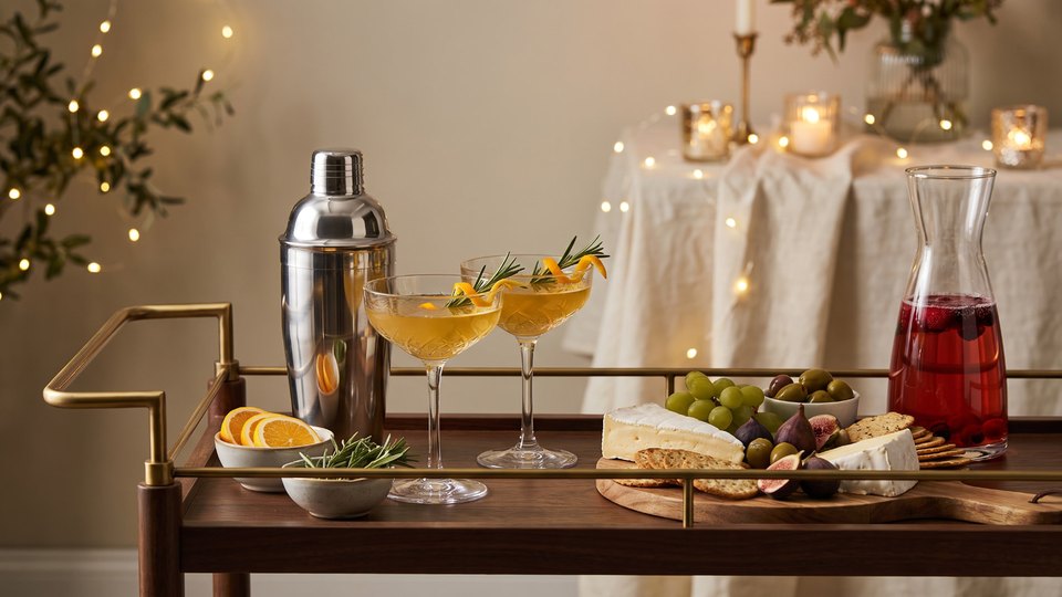 Holiday celebrating essentials: cocktail shaker, coupe glasses with rosemary and orange twists, a carafe of berry punch, and a board of brie, grapes, figs, and crackers