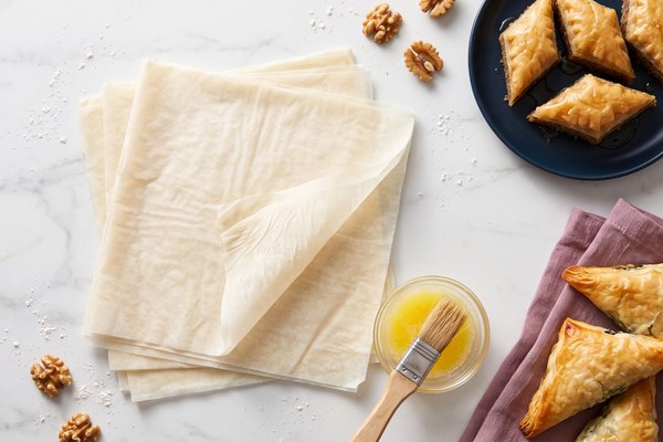 Flat lay of raw phyllo dough sheets, baklava, and spanakopita with melted butter and a pastry brush