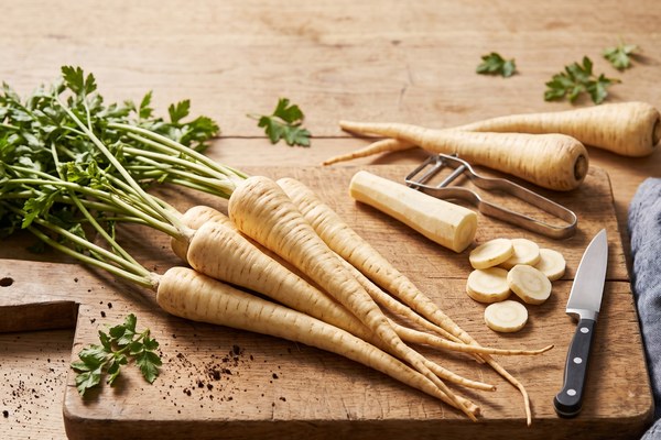 Fresh whole and sliced parsnips on a wooden cutting board with a peeler and knife.