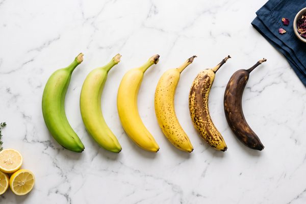 Six bananas on a white marble surface arranged from green to yellow to heavily spotted brown, showing each stage of ripening