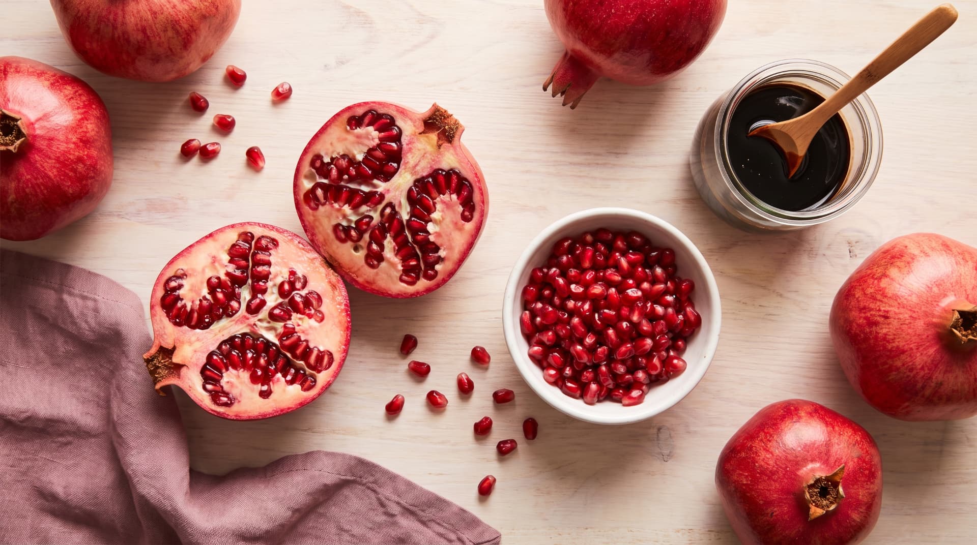 Overhead flat lay of a halved pomegranate showing glossy red seeds, a bowl of loose pomegranate arils, whole pomegranates, and a small jar of pomegranate molasses on a light wood surface with a mauve linen napkin.