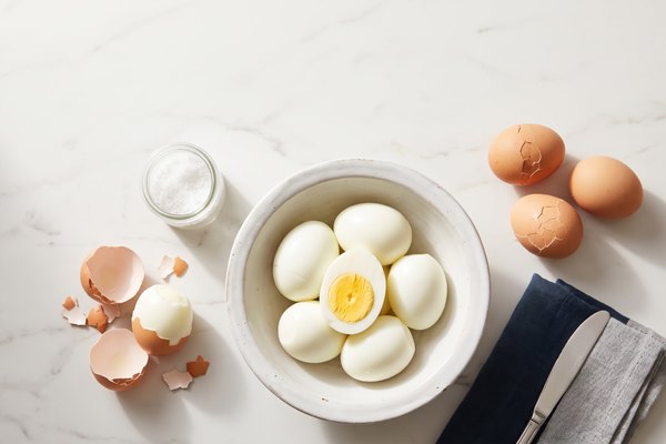 A bowl of peeled hard boiled eggs next to shells being peeled away, illustrating hard boiled egg storage and peeling
