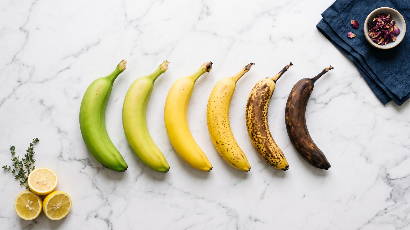 Six bananas on a white marble surface arranged from green to yellow to heavily spotted brown, showing each stage of ripening